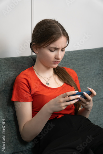 Worried young girl is reading upsetting news on her smartphone while sitting on a gray sofa in her living room, experiencing a moment of concern and uncertainty. Vertical photo