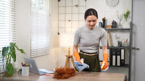 An Asian woman wearing orange rubber gloves wipes a wooden desk with a microfiber cloth and spray bottle in a modern home office, focusing on cleanliness, hygiene, and workspace maintenance.