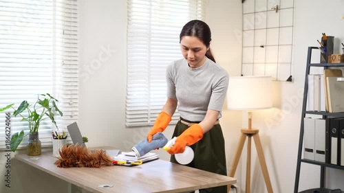 An Asian woman wearing orange rubber gloves wipes a wooden desk with a microfiber cloth and spray bottle in a modern home office, focusing on cleanliness, hygiene, and workspace maintenance.