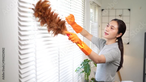 An Asian woman wearing orange rubber gloves cleans white window blinds with a microfiber cloth in a modern, bright home interior, focusing on hygiene and household maintenance.