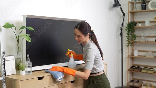 A young woman wearing protective gloves cleans a wooden cabinet and television screen with a spray bottle and cloth, representing home cleaning, housekeeping, and household hygiene in a modern living 