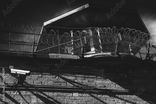 Exterior prison guard tower and wall harshly illuminated by floodlights at night, barbed wire, surveillance camera, nobody