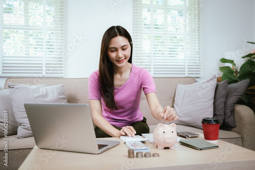 Young Asian woman sitting on a sofa using a laptop and taking notes while managing personal finances at home, with a piggy bank, cash, coins, and coffee representing budgeting and saving.