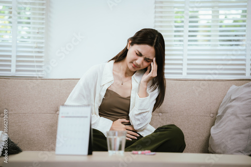 Young Asian woman sitting on a sofa holding her lower abdomen in pain, experiencing menstrual cramps or stomach ache at home, with a calendar and medicine symbolizing period health issues.