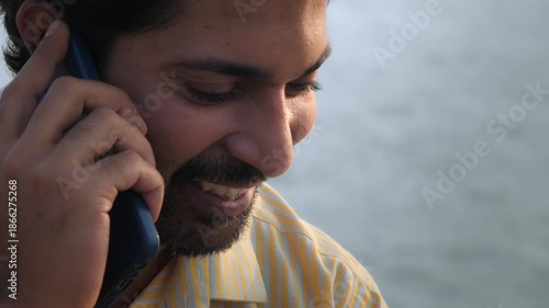 A daylight outdoor close up shot of a happy Indian Asian middle aged bearded man or male smiling and talking on a mobile phone or smartphone with a water body in the background or sea on a sunny day.