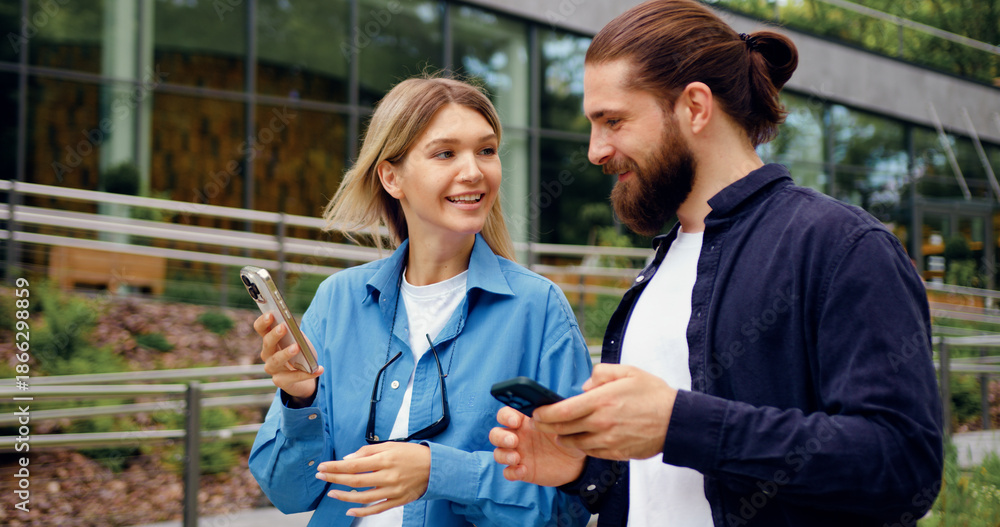 Obraz premium Two young adults man and woman strolling in a park while checking their mobile phones, sharing something on the screen and smiling during a friendly conversation
