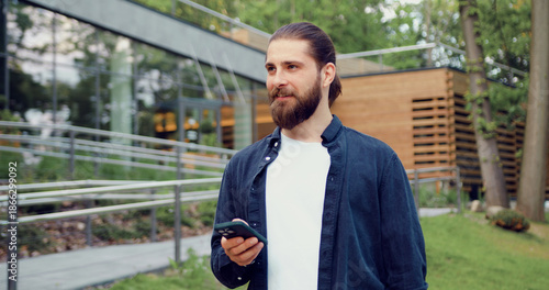 Wallpaper Mural Portrait of a handsome bearded man looking at his smartphone screen in a city park, thoughtfully calculating, outdoors in an urban environment. Torontodigital.ca