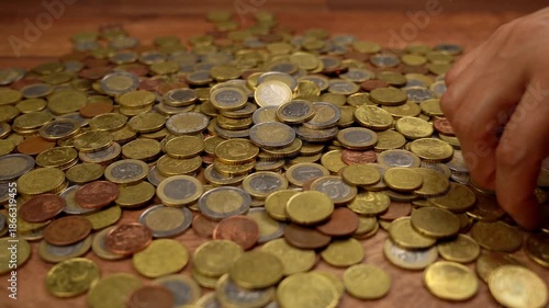 Close-up of a person's hand sorting through a large pile of various euro coins on a wooden table, counting and stacking the money