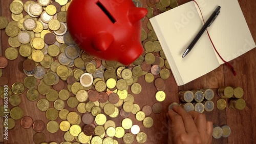 Overhead view of hands counting and stacking euro coins on a wooden table. A red piggy bank and a notepad represent saving and accounting