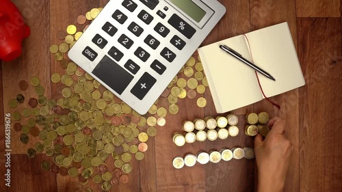 Overhead view of a person managing finances, stacking euro coins next to a calculator, a notepad, and a piggy bank on a wooden surface
