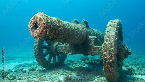 Ancient cannon covered in coral and marine growth rests on a rocky seabed in clear blue ocean water, depicting a sunken shipwreck artifact and underwater historical relic