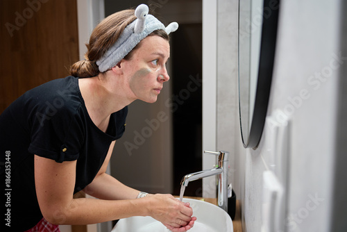 Woman standing at bathroom sink washing face while wearing clay face mask and headband