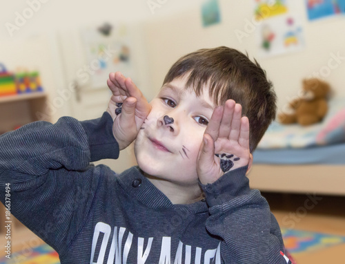 Close-up portrait of a little boy with cat face painting posing in a playroom with toys on the background.