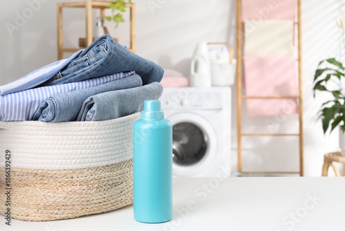 Laundry basket with clothes and bottle of detergent on white table indoors. Space for text
