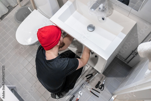 Handyman installing a new faucet and plumbing lines under a bathroom sink. Tools on a tiled floor in a modern bathroom illustrate home renovation, repair, maintenance, and proper installation work