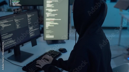 Hacker wearing black hoodie coding on multiple monitors in dark room modern office during night shift, cybersecurity risk, computer hacking, data breach, digital crime, network security, technology.