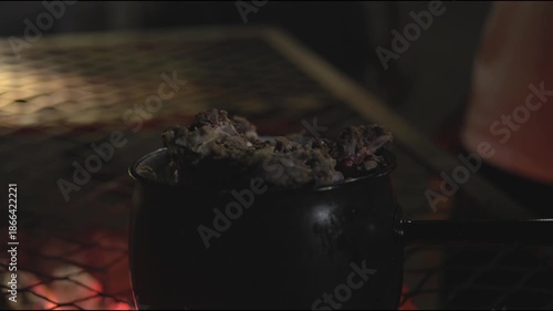 Night time cooking, grilling, and warming fresh goat meat over fire and pot on grill top grate in Africa with Zimbabwe culture, cooks, and people working together to feast on red meats outdoors