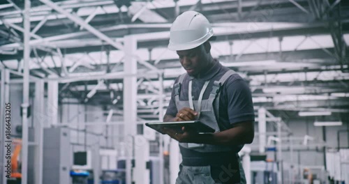 Black Male Technician Wearing Safety Helmet Controls Production Processes With Digital Controller in High Tech Factory Filled With Robotic Arms. Concept of Industry 4.0 and Automation Robotic Factory.
