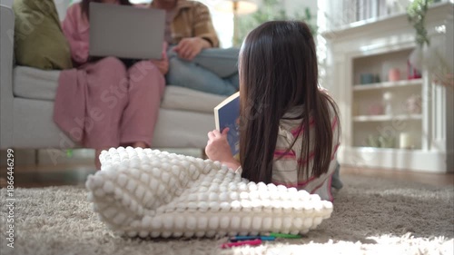 A young girl lying on a soft carpet and drawing at home while her parents relax on the sofa in the background