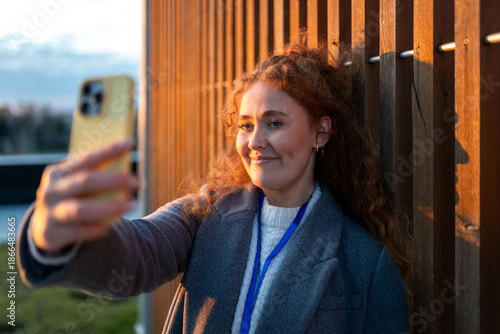 Obraz na plátně Woman with red curly hair taking selfie on smartphone during golden hour outdoor