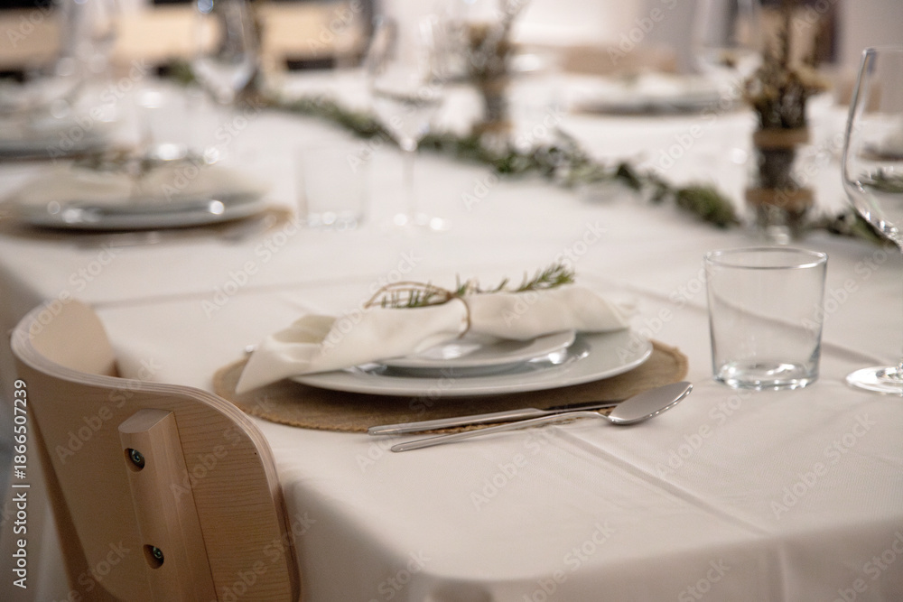 Fototapeta premium A neatly arranged dining table shows a single place setting with white plates, cutlery and a cloth napkin tied with twine and fresh rosemary, prepared for an elegant family dinner in a cozy home.