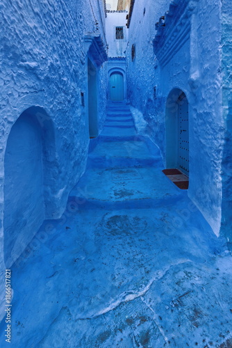 Narrow, stepped, sloped pedestrian alley painted in shades of blue -maybe the bluest in town- leading to a closed wooden door. Chefchaouen-Morocco-033