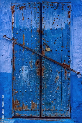 Chipped, dilapidated, studded old wooden door closed with a security bar in the medina, in various paint-faded shades of blue. Chefchaouen-Morocco-034