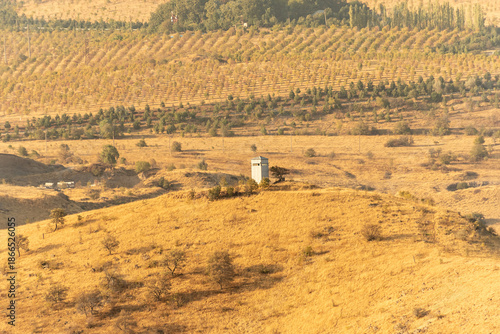 A tall tower stands alone on a hillside. Surrounding it, fields stretch across the landscape with rows of trees in the distance. The sun casts warm light over the area.