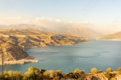 A landscape shows hills and mountains surrounding a river. The sun shines on the scene during late afternoon. Trees grow near the water, and the sky has few clouds.
