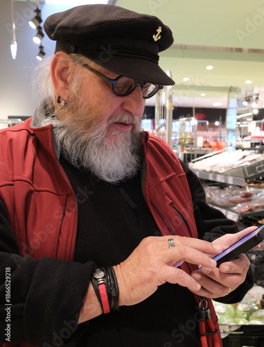 Senior man with a beard and glasses, wearing a black cap with an anchor emblem and red vest, is looking at a phone in a grocery store. The man is  checking his shopping list