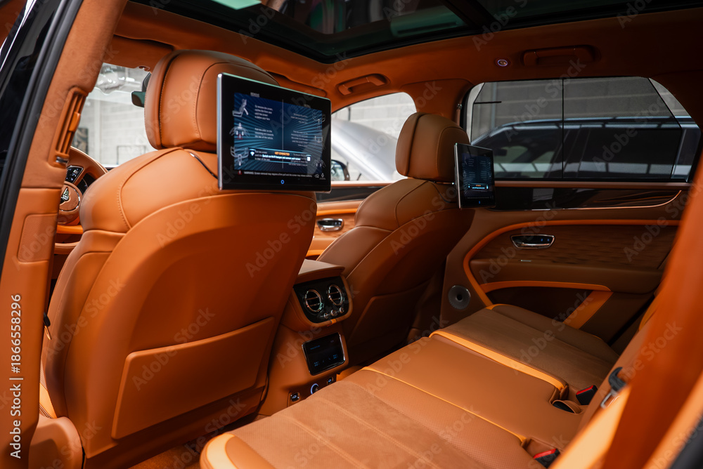 Obraz premium Rear cabin of a Bentley Bentayga Speed in an indoor bay shows tan leather, diamond quilted doors, dual rear screens, panoramic roof, and climate console in soft light.