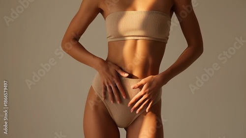 Slim woman caressing her flat stomach in warm sunlight shadows, wearing beige lingerie, showcasing healthy skin and weight loss concepts.