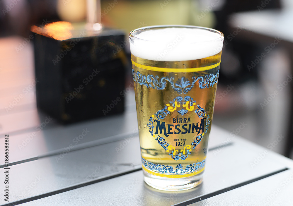 Fototapeta premium SIENA, ITALY – OCTOBER 7, 2025: Sicilian lager 'Birra Messina' beer in decorated glass. The beer was founded in 1923 and is part of the Heineken group since 1988. Selective focus