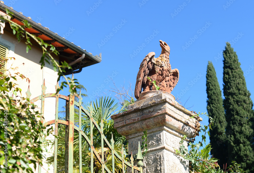 Fototapeta premium Ancient sculpture of an eagle on stone socket at iron gate with clear blue sky on an october day in Poggibonsi, Tuscany