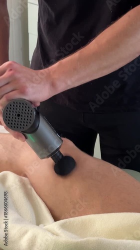 A massage therapist uses a massage gun to treat a client’s leg muscles, applying pressure with controlled motion