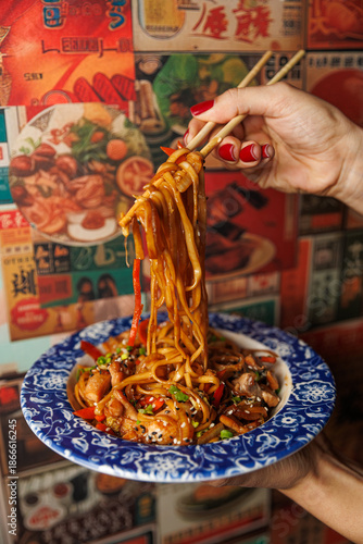 Hand holding chopsticks with noodles and vegetables in a decorative bowl against a colorful food-themed background