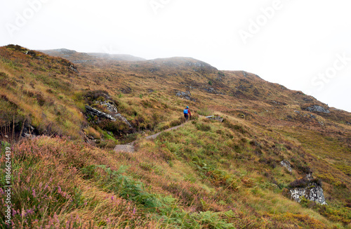 Tiny backpacker figure climbing up a vast curving trail in the Scottish wilderness on a misty autumn day