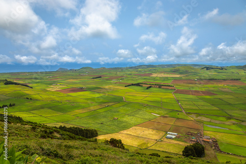 Panoramic view of the colorful agricultural fields from Serra do Cume lookout on Terceira Island, Azores, Portugal. Lush green, yellow, and brown farmlands form a scenic patchwork landscape 