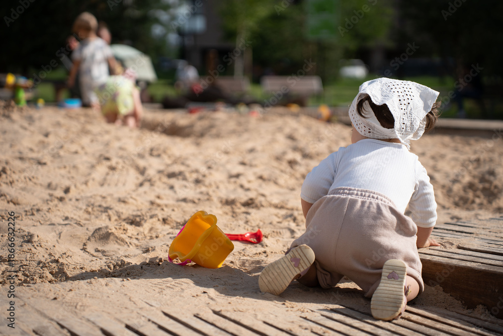 Obraz premium a toddler in a white bonnet crawls in a sunlit sandbox with a yellow bucket and red shovel, surrounded by blurred children playing in a lively park setting, leisure, relax, carefree, day, lively