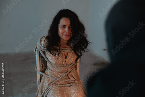 Woman sitting on a chair and tied with a rope, crying and looking at her captor in basement. Crime, kidnapping, violence concept