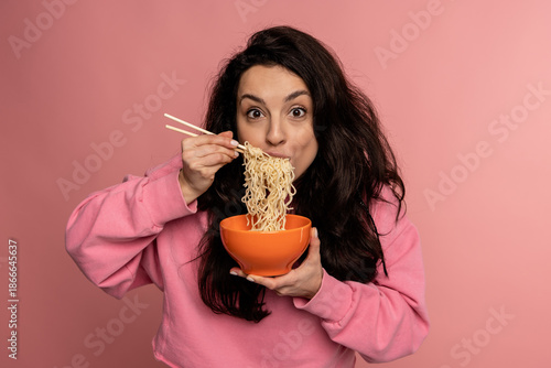 Hungry dark-haired young Caucasian woman eating pasta with an appetite during the studio photo shoot on the pink background. Japanese food concept