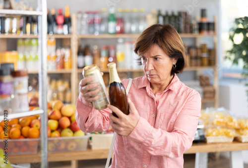 Bild auf Leinwand Concerned old woman deciding between kinds of beer in the supermarket