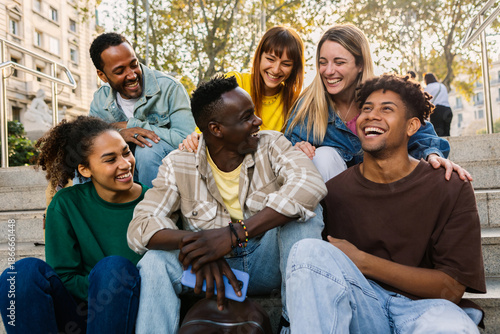 Cheerful millennial group of diverse friends sitting outdoors on city stairs, talking and laughing while enjoying free time in an urban environment. Youth community and friendship lifestyle concept. #1866661448