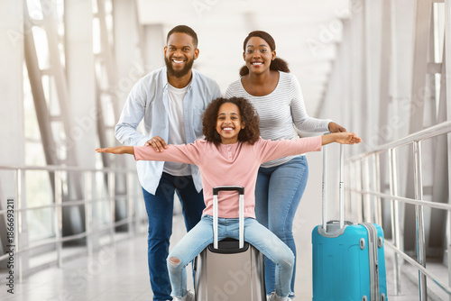 Wallpaper Mural Happy Loving Family In Airport. Portrait of joyful excited black girl sitting on suitcase and laughing, spreading arms imitating plane, smiling cheerful parents posing, standing in terminal hall Torontodigital.ca