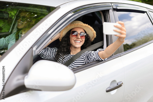 Pretty middle-eastern millennial woman in sunglasses and summer hat enjoying car journey with husband or boyfriend, outstretching hand through auto window and taking selfie, using smartphone