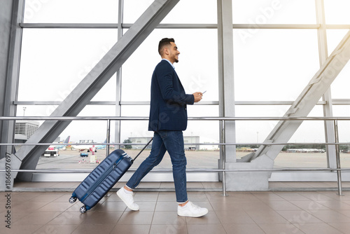 Obraz na plátně Young middle-eastern businessman with suitcase and smartphone in hands walking i