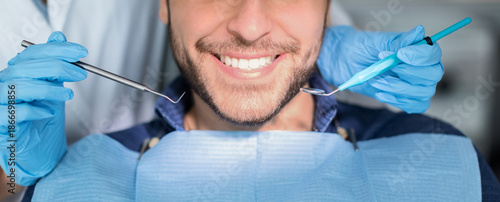 Fotografija Unrecognizable male patient having teeth whitening procedure at modern dental clinic, smiling at camera, dentist hands in rubber gloves with dental tools