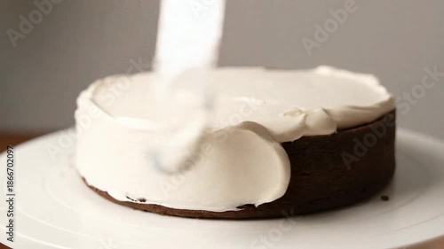 A close-up shot of a dark chocolate cake being frosted with smooth white cream using a spatula on a white cake stand.