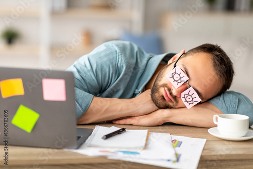 Lazy unproductive young guy wearing funny sticky notes with open eyes on his glasses, sleeping at workplace. Tired overworking millennial man falling asleep at home office