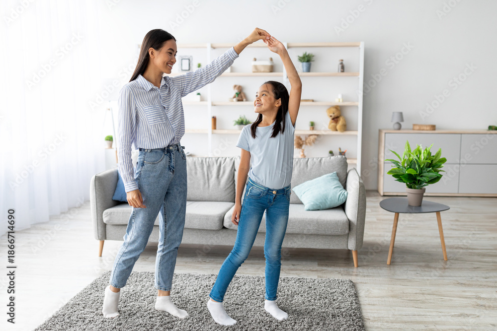 Fototapeta premium Cheerful asian mother and daughter dancing to music, holding hands and enjoying spending time together, having fun in living room interior at home. Full length shot